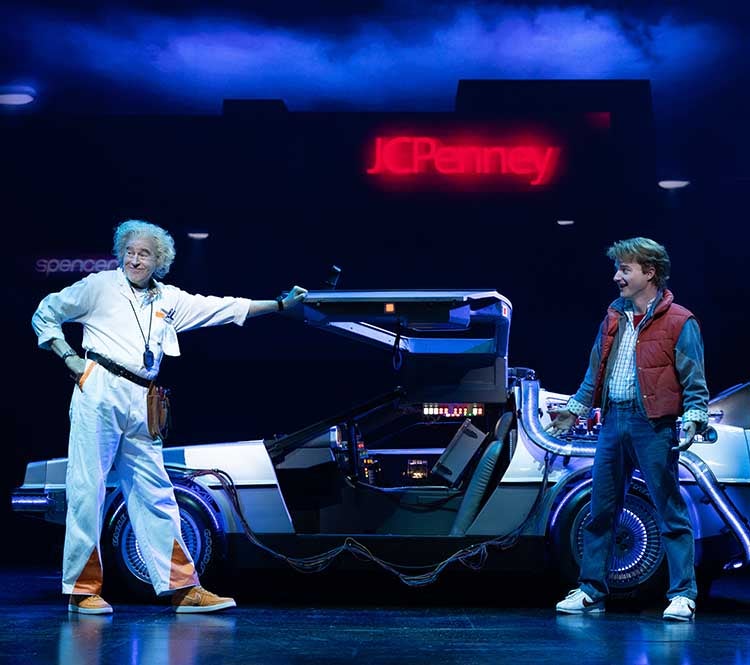 Back To The Future: Don Stephenson as Doc Brown and Lucas Hallauer as Marty McFly share a pivotal moment in the touring production of Back to the Future: The Musical, standing beside the iconic DeLorean under a futuristic glow.