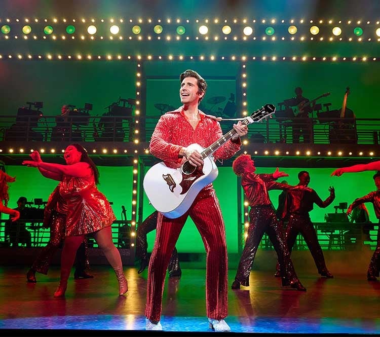 Nick Fradiani as Neil Diamond holds a white acoustic guitar while wearing a red sequin outfit. Dancers appear on stage behind him.