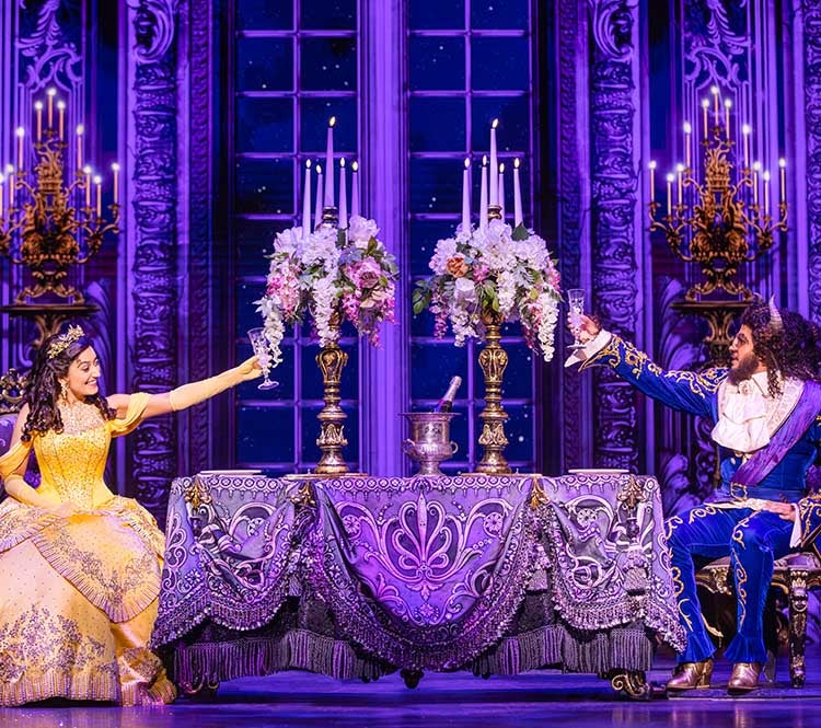 Beauty and the Beast: Belle, dressed in a yellow ballgown sits at a table across from the Beast dressed in a blue suit, each holding a glass, arms raised. Two flower arrangements decorate the table with candles.