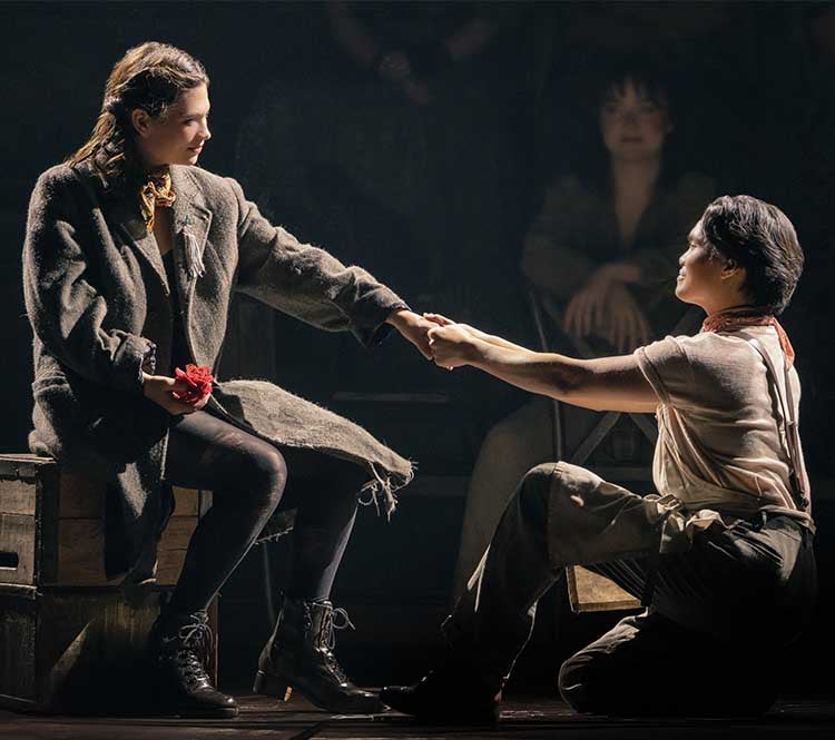 Hadestown: Orpheus and Eurydice sit facing each other. Orpheus is holding her left hand with both of his. In Eurydice’s right hand is a red carnation.