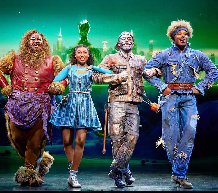 The Wiz: The Lion, Dorthy, the Tinman, and the Scarecrow walk arm in arm smiling. The Emerald City is shown behind them.